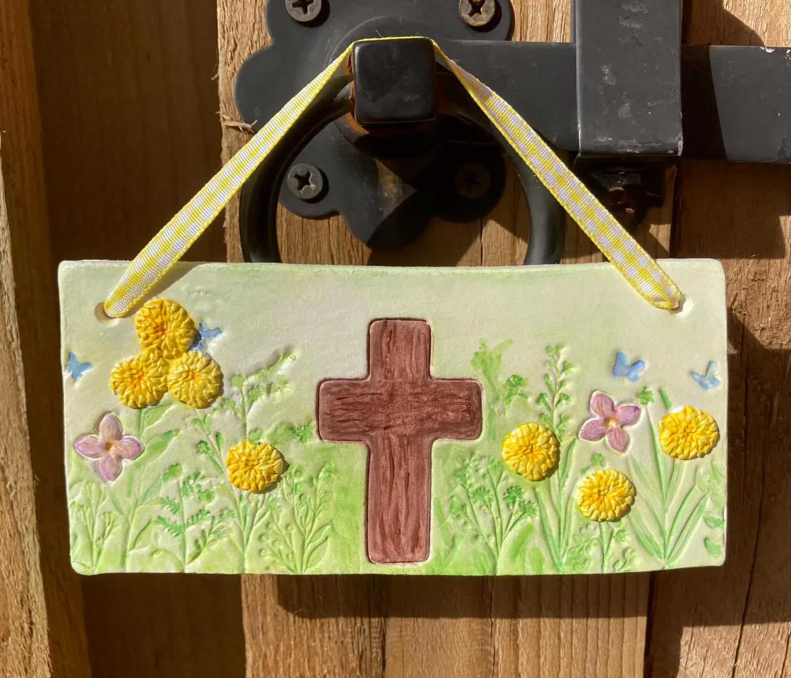 A long air-dry clay plaque with a brown cross among yellow and pink flowers and blue butterflies, hanging from a gingham ribbon on a gate latch