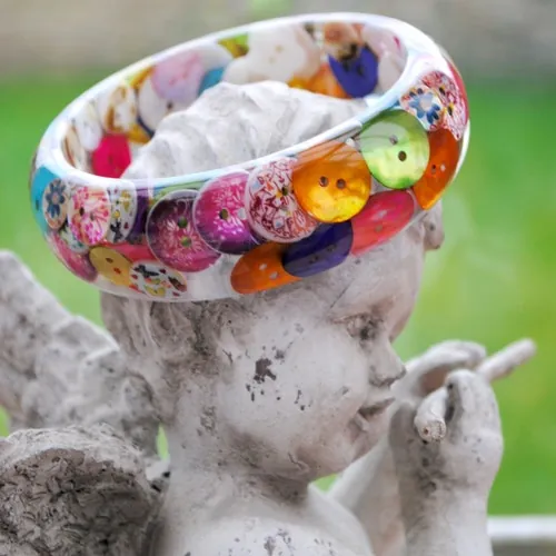A clear resin bangle filled with colourful buttons, resting on the head of a small stone cherub in a garden